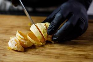 Person with black gloves cuts peeled orange on wooden cutting board into pieces photo