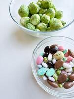 Brussels sprouts in clear glass bowl above assorted candy in another bowl on white surface. Healthy eating concept. photo