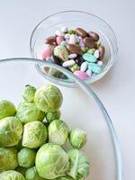 Brussels sprouts in clear glass bowl above assorted candy in another bowl on white surface. Healthy eating concept. photo