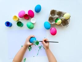 Childrens hands paint Easter eggs with brush surrounded by colorful eggs and jars of paints on white table, creative holiday activity. photo