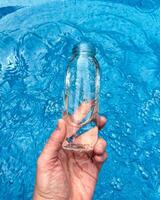 Hand holding clear glass bottle partially filled with water against sparkling blue water background. Clean water concept. photo