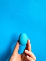 Hand balancing blue egg on a fingertip against solid blue background, minimalist concept for balance, Easter, and simplicity with space for text. photo