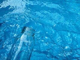 Clear glass bottle partially submerged in rippling blue water with reflections and refractions, top view. Clean water concept. photo