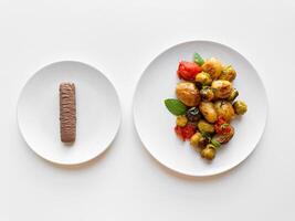 Two white plates on table, one with single chocolate bar and other with roasted baby potatoes and Brussels sprouts, contrasting healthy and indulgent food choices. photo