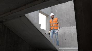 Builder repairman, foreman in protective helmet and vest stands at workplace in building and holds ruler and toolbox in hands video