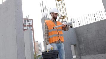 Builder repairman, foreman in protective helmet and vest stands at workplace in building and holds ruler and toolbox in hands video