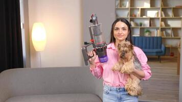 un linda joven mujer es participación un sin cable vacío limpiador con un sofá adjunto archivo en uno mano y un perro en el otro mano video