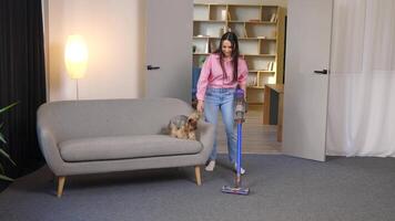 un linda joven mujer es Pasar la aspiradora a hogar con un sin cable vacío limpiador y acariciando su linda pequeño perro con su mano video