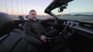 A man sits in a luxurious convertible, Mature man with a content expression driving a convertible car at sunset. video