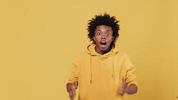 Young black guy looks into the distance with a puzzled face, then finally recognizes something and becomes extremely overjoyed with raising up his hands on a yellow background in a studio video