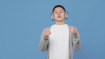 Jeune asiatique homme dans écouteurs méditer sur une bleu Contexte dans une studio. conscient, calme et détendu gars de liaison le pouce et index sur tous les deux mains, en mettant leur à le côtés video