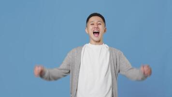 Young asian guy carefully looking into the distance, and then becomes happy and joyful and raising his hands up on a blue background in a studio video