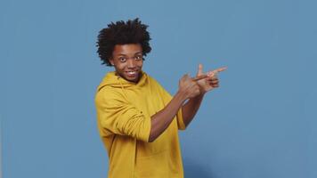 Young smiling black guy pointing with both hands to the camera and then to the copy space on a blue background in the studio. Concept of advice and promotion video