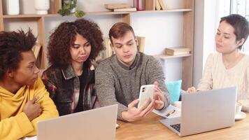 Young man demonstrating on the smartphone the concept of his work ideas to multiracial corporate employees team while sitting together in office on the common table video