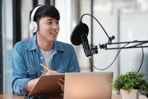 A man wearing headphones is sitting at a desk with a laptop and a notepad photo