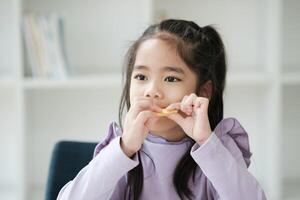 un joven niña es comiendo un pedazo de comida mientras sentado en un habitación con estantería foto