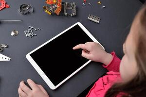 pequeño niña usos un tableta en un escritorio, rodeado por pequeño partes y tornillos. el concepto de educación y composición de asignaturas, programación para niños foto