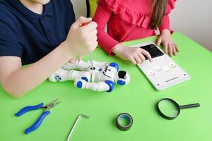 Children's hands assemble robot on workbench, screwing parts while reviewing programming code on tablet. Concept of learning and hands-on education photo