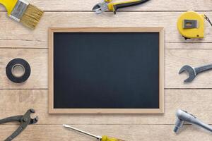 Clean chalkboard surrounded by tools, brush, pliers, screwdriver, tape measure, adhesive tape, wrench on wooden table. Perfect for text, logo promotion. Top view, flat lay composition. DIY concept photo