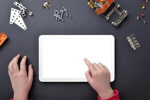 Tablet mockup on work desk. Child's hands touch isolated display. Top view, flat lay desk composition with small metal parts and screws photo