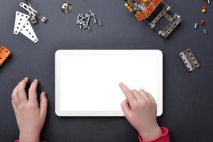 Tablet mockup on work desk. Child's hands touch isolated display. Top view, flat lay desk composition with small metal parts and screws photo