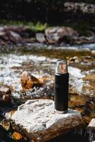 Thermo bottle to keep the heat inside, thermos with coffee standing on a rock in the stream, mountain stream, metal vessel for liquid, tourist thermos on a hike photo