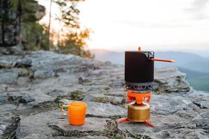 Camping kitchen in the mountains against the backdrop of sunset, boil water on a tourist burner, brew tea, a mug and a thermos, a camping pot, a tourist's pot. photo