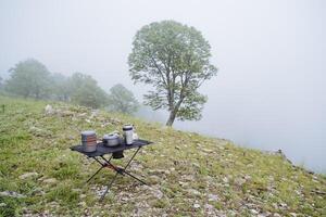 Table stands on rocks in the forest foggy weather camping furniture tree in the cloud cloudy weather food in a pot camping kitchen set of appliances photo