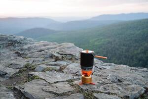 A gas burner with a tourist pot stands against the backdrop of mountains on a hike, boiling water in a pot, a cooking system in the wild. photo