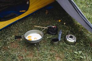 Breakfast in a tent in nature, fried glaze in a frying pan stands on the grass under the canopy of the tent, a gas burner, a small kettle on a hike, a climber's food. photo