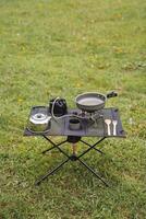 Picnic in nature at the table, tourist utensils for camping, equipment for cooking in the forest, gas burner, kettle with mug, frying pan. photo