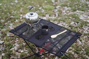 Camping furniture folding table made of netting, camping utensils, a glass of tea, a metal kettle stands on a gas burner. photo