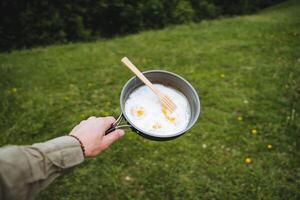 A hand holds a frying pan with fried eggs on fire, icing on a hike, a tourist's food on a hike through the forest. wooden fork. photo