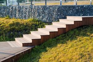 Steps from the boards descend down, a wooden staircase in the park, a path for walking, a place of rest for citizens, in the morning grass in the rays of the sun. photo