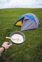 Fried scrambled eggs in a frying pan, the hand holds against the background of the tent, a camping breakfast in nature, the morning food of an extreme tourist, camping in the field. photo