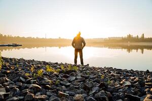 A guy on the lake stands by the water in the rays of sunlight, meditation in the morning at dawn, sun glare, quiet surface of the lake, city recreation park, Zen photo