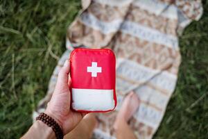 un parte superior ver de un primero ayuda equipo en naturaleza, un excursionismo bolso con medicamentos, un persona sostiene un rojo primero ayuda equipo en su mano. foto