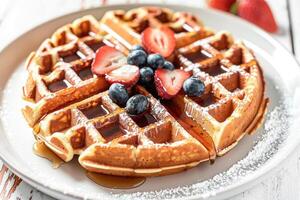 Waffles drizzled with syrup and adorned with fresh strawberries, blueberries on white plate on white wooden table. Tempting breakfast or dessert option, promoting culinary, brunch menus photo