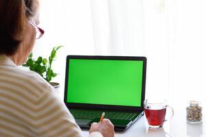 Over shoulder view of senior woman in glasses using laptop with green screen. Perfect for illustrating technology adoption among older adults, remote work, and digital literacy photo