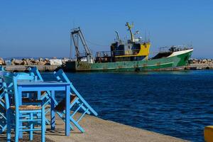 Blue wooden table and chair of a typical fish tavern at the port. Fishing ship on the background. Aegean island Chios in Greece on an autumn day. Greek holidays and destinations photo