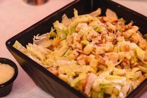 Close-up of large portion of salad with chicken, lettuce, cabbage, croutons, cheese and sauce served in the black bowl on white background in greek tavern. Table setting. Mediterranean cuisine photo