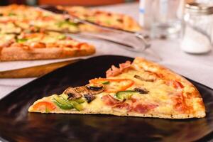 Close-up a piece of large delicious pizza with mushrooms, piper, cheese, ham served on the black plate outside in greek tavern. Table setting. Customer service. Mediterranean cuisine photo