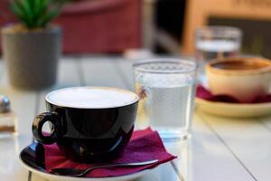 Cup of cappuccino coffee in black cup, spoon on saucer, cup of greek coffee with glass of water on a wooden white table at the cafe. Selective focus photo