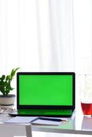 Concept of distance education, Vertical view of laptop with green screen, notebook, pen, glasses, cup of tea, flower on the table in front of window. Selective focus. photo