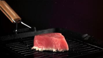 Super schleppend Bewegung Thunfisch Steak ist gebraten im ein Grill schwenken mit spritzt von Öl. auf ein schwarz Hintergrund.gefilmt auf ein schnelle Geschwindigkeit Kamera beim 1000 fps. video