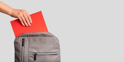 Hand putting red book, textbook into backpack, school bag. Banner background photo