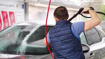 Young man washes his car at a self-service car wash using a hose with pressurized water and foam. High quality 4k footage video