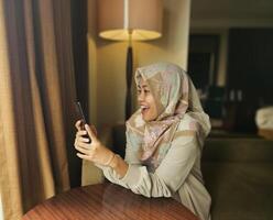 contento asiático musulmán mujer vistiendo hijab sonriente cuando leyendo texto mensaje o charla en su teléfono con difuminar fondo, comunicación en teléfono, moderno global Internet social medios de comunicación concepto foto