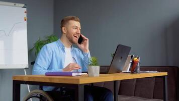 motivée de bonne humeur handicapé pigiste parlant sur le téléphone et prise Remarques séance dans fauteuil roulant à bureau. loin travail et invalidité concept video