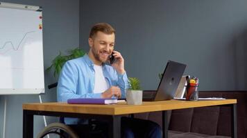 Motivated cheerful handicapped freelancer talking on the phone and taking notes sitting in wheelchair at office. Distant work and disability concept video